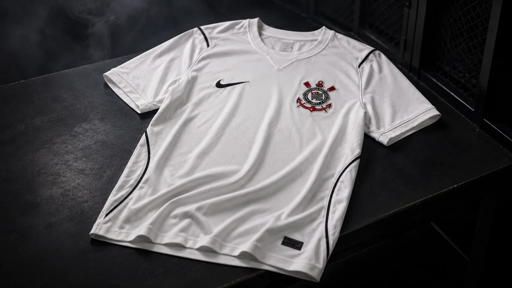 Camisa branca do Corinthians apoiada sobre uma superfície escura, em ambiente de vestiário com iluminação dramática, fumaça ao fundo e detalhes do uniforme em destaque.