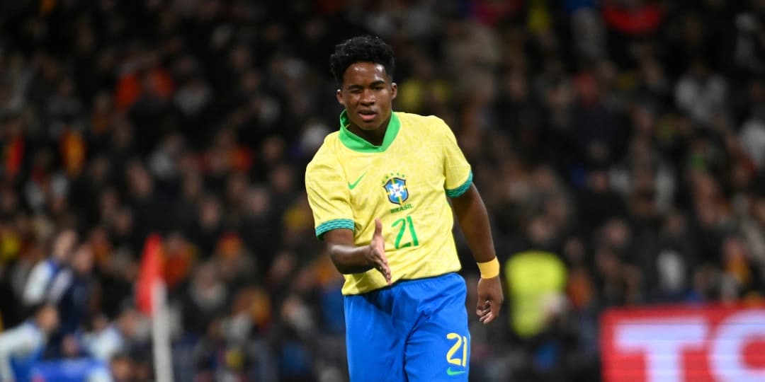Fotografia do jogador de futebol Endrick em campo, vestindo o uniforme da Seleção Brasileira com a camisa amarela número 21 e calções azuis. Ele aparece estendendo a mão direita em um gesto no gramado durante uma partida à noite, com o público na arquibancada ao fundo. A imagem ilustra o contexto dos bastidores da quase contratação de Endrick no Corinthians antes de sua projeção mundial.