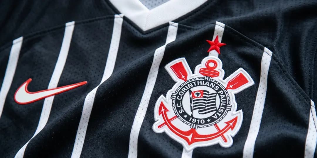 Close-up detalhado da camisa oficial do Sport Club Corinthians Paulista, destacando o escudo bordado com âncora e remos sobre o tecido preto com listras verticais brancas. A imagem representa a identidade visual do time, usada nos confrontos contra o maior rival do Corinthians.