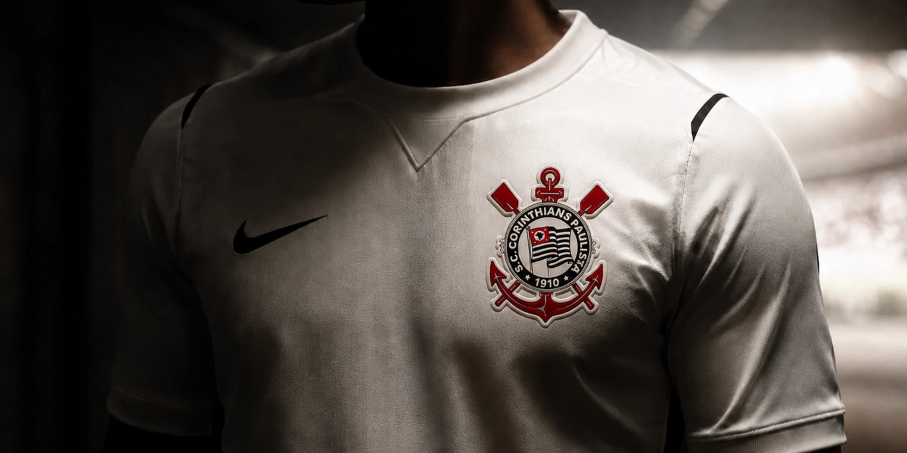 Close-up editorial de uma pessoa em silhueta usando camisa branca do Corinthians, com o escudo corrigido em destaque no peito, em ambiente escuro de túnel de estádio, ilustrando o tema quem o Corinthians vai contratar na janela do meio do ano.